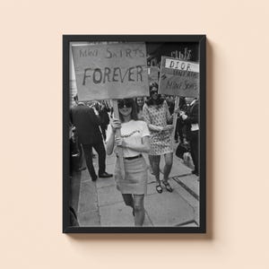 May include: Framed black and white photograph of a protest. Women are holding signs with the slogans "Mini Skirts Forever" and "Dior Unfair Mini Skirts". The women are wearing mini skirts and dresses.
