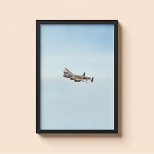 Pode incluir: Impressão emoldurada de um bombardeiro vintage voando contra um céu azul claro. A aeronave, predominantemente verde-oliva e marrom, exibe hélices e marcações. A impressão está dentro de uma moldura preta.