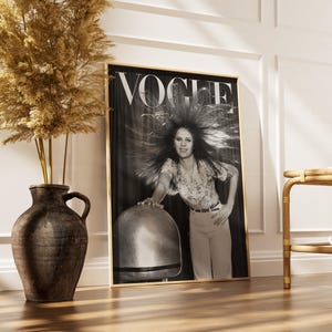 Puede incluir: Una portada de revista Vogue en blanco y negro enmarcada, con una mujer con cabello voluminoso. La revista est&aacute; en un marco dorado, apoyada contra una pared blanca. A la izquierda hay un jarr&oacute;n marr&oacute;n grande con plantas secas y a la derecha una mesa auxiliar de bamb&uacute;.