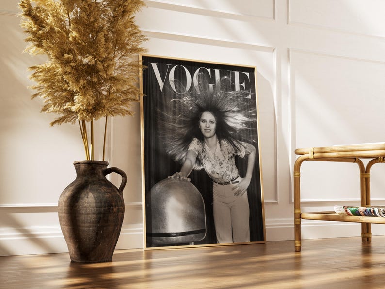 Puede incluir: Una portada de revista Vogue en blanco y negro enmarcada, con una mujer con cabello voluminoso. La revista est&aacute; en un marco dorado, apoyada contra una pared blanca. A la izquierda hay un jarr&oacute;n marr&oacute;n grande con plantas secas y a la derecha una mesa auxiliar de bamb&uacute;.