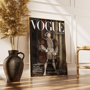May include: A framed print of a magazine cover titled "ANCIENT VOGUE 1540 AD" featuring a portrait of King Henry VII of England. The print is in a gold frame and is propped against a white wall. A large brown vase with dried plants is to the left.
