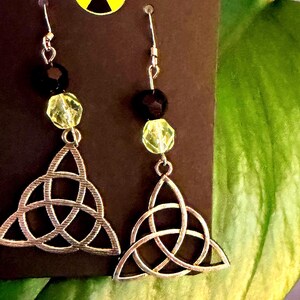 May include: Silver-toned Celtic knot earrings with black and yellow beads. The earrings hang from silver hooks and feature a triquetra design. The background includes a dark brown card and green foliage.