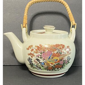 May include: A cream-colored ceramic teapot featuring a vibrant, multi-colored floral and bird motif on the body. The teapot includes a matching lid with a small knob and a curved, light-brown woven wicker handle attached to two side loops.
