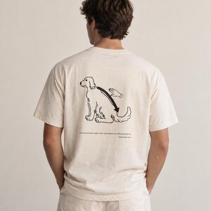 May include: A cream-coloured short-sleeve t-shirt featuring a black line drawing on the back of a dog being petted. Below the illustration, the text reads: THE RIGHTEOUS CARE FOR THE NEEDS OF THEIR ANIMALS. PROVERBS 12:10. The shirt has a crew neckline.