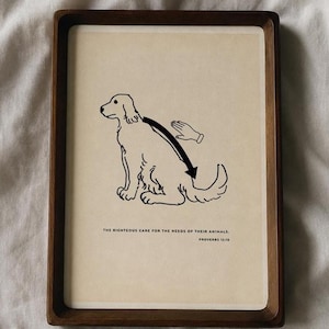 May include: A rectangular wooden tray containing a cream-coloured print of a dog being petted by a hand. The black line drawing includes an arrow indicating a petting motion along the dog's back. Text reads: THE RIGHTEOUS CARE FOR THE NEEDS OF THEIR ANIMALS. PROVERBS 12:10.