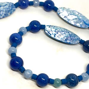 May include: A necklace featuring a repeating pattern of round dark blue beads, smaller light blue beads, and elongated oval blue shell-like components with a cracked texture, all separated by small, flat, cylindrical blue spacers.