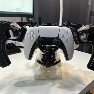 Peut inclure: Une manette PlayStation 5 blanche et noire avec une fixation de poignée noire personnalisée. La manette est montée sur un support blanc. L'ensemble est conçu pour un contrôle et un confort de jeu améliorés.