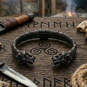 May include: A dark metallic open-ended bracelet featuring two sculpted animal heads at the terminals, resting on a wooden surface engraved with runic symbols, accompanied by two knives with wooden handles and several small carved stone blocks.