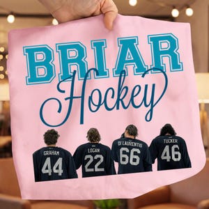 Può includere: Una borsa tote rosa con la scritta "BRIAR Hockey" in turchese. Sotto, le spalle di quattro giocatori di hockey con maglie blu navy con numeri e nomi bianchi: Graham, Logan, Di Laurentis e Tucker.