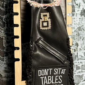 May include: Black leather tie with a decorative chain and a wooden display. The tie features a patch with the text "DON'T SIT AT TABLES THAT JESUS WOULD HAVE FLIPPED". Embellished with rhinestones and a floral accent.