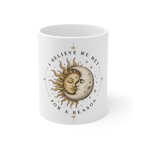 May include: White ceramic mug featuring a celestial design with a sun and moon embracing, surrounded by stars. The mug has the text "I BELIEVE WE MET FOR A REASON" in a circular pattern around the design.