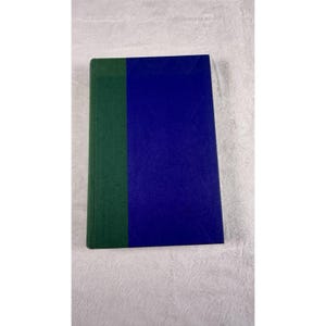 May include: A hardcover book with a dark green spine and a blue cover. The book is rectangular and appears to be closed. The book is lying on a white, textured surface.