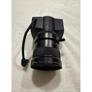 Könnte beinhalten: Ein schwarzes TV-Zoomobjektiv mit einer Brennweite von 9,5-114 mm. Das Objektiv hat einen geriffelten Fokusring und ein Kabel, das sich von oben erstreckt. Markierungen zeigen die Entfernung in Fuß und Metern an.