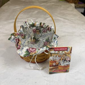 May include: A round woven wooden basket with a single arched handle and a ruffled fabric liner featuring a floral pattern of pink roses and green leaves. A clear glass insert sits inside, and a product card labelled 1998 Mother's Day Basket is included.