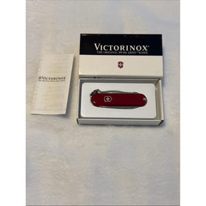 Puede incluir: Una navaja suiza Victorinox roja en su embalaje original. La caja es blanca con la parte superior negra y el nombre de la marca. Un pequeño folleto de instrucciones está a la izquierda de la caja.