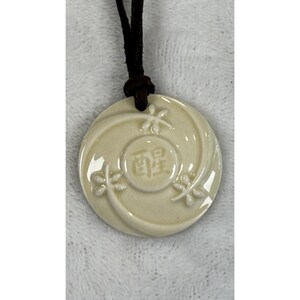 May include: Cream ceramic pendant with embossed floral designs and Japanese characters. The round pendant is attached to a dark brown cord. The pendant has a glossy finish and is centered in the frame.