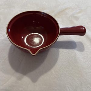May include: A burgundy ceramic sauce bowl with a handle. The bowl has a white rim and a spout for easy pouring. The handle is the same burgundy colour as the bowl. The bowl is on a white surface.