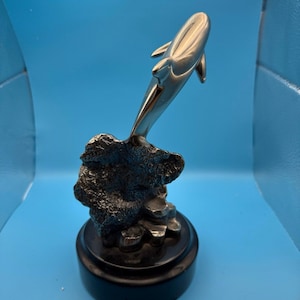 May include: A silver dolphin sculpture leaping from a textured, dark gray rock formation. The sculpture is mounted on a black, circular base, set against a blue background. The dolphin's sleek form contrasts with the rough texture of the rock.