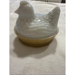 Puede incluir: Un plato blanco con forma de gallina con tapa, sobre una base dorada. La gallina tiene texturas detalladas de plumas y un pico prominente. El plato es probablemente de cerámica o vidrio y está diseñado para servir o guardar.