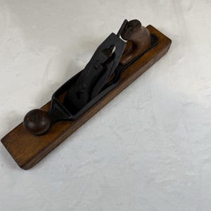 May include: A vintage wood-bodied hand plane featuring a long rectangular wooden base, a black metal blade assembly, and a rounded wooden handle at the rear. The tool includes a front wooden knob and a central adjustment mechanism for the metal blade.