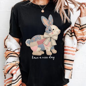 May include: Black t-shirt featuring a patchwork bunny design with floral and gingham patterns. The bunny has a large bow and the words "have a nice day" are printed below the bunny.