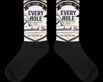 Puede incluir: Un par de calcetines blancos con talones y punteras negras, con un diseño gráfico de palos de golf y el texto "EST. PLANO TX", "EVERY HOLE IS A comeback Story" y "PAR FOR THE WIT" impreso en azul marino, rojo y dorado en la caña.