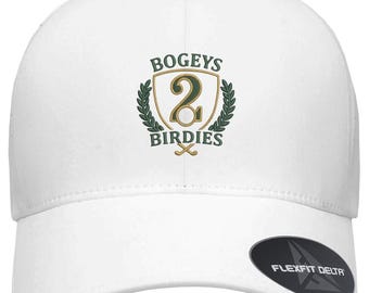 Puede incluir: Una gorra de béisbol blanca con un logotipo bordado que incluye el texto BOGEYS y BIRDIES alrededor de un escudo con el número 2 y dos palos de golf cruzados. Una pegatina ovalada negra en la visera muestra el texto FLEXFIT DELTA ADJUSTABLE.