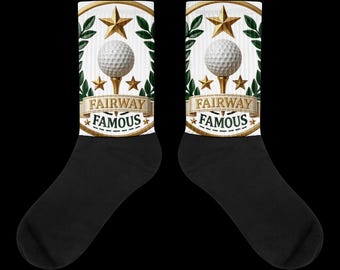 Puede incluir: Un par de calcetines negros con un gráfico blanco en la parte superior. El gráfico muestra una pelota de golf sobre un tee, rodeada de estrellas doradas y hojas de laurel verdes, con una cinta que contiene el texto FAIRWAY FAMOUS en oro y negro.