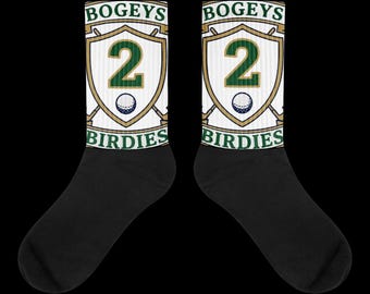 Puede incluir: Un par de calcetines negros con un panel gráfico blanco en la pantorrilla. El diseño incluye un escudo con palos de golf cruzados, el número 2 en verde, una pequeña pelota de golf y el texto BOGEYS BIRDIES en letras verdes sobre fondo blanco.