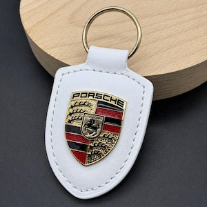 May include: White leather keychain with a gold-colored ring. The keychain features the Porsche emblem in gold, red, black, and yellow. The emblem is stitched onto the white leather.