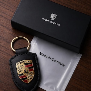 May include: Black leather Porsche keychain with gold ring and emblem, presented with a black box and a packet that reads "Made in Germany". The keychain features the Porsche logo in gold, red, and black.