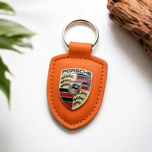 May include: Orange leather keychain with a silver key ring. The keychain features the Porsche emblem in gold, black, red, and silver. The keychain is shield-shaped and has stitched edges.