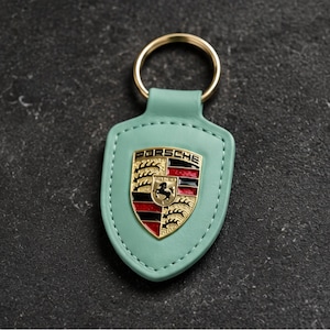 May include: A mint green shield-shaped keychain featuring a gold-tone metal Porsche crest emblem in the center. The crest displays black, red, and gold enamel details with the word PORSCHE printed at the top. A gold-tone metal ring is attached.