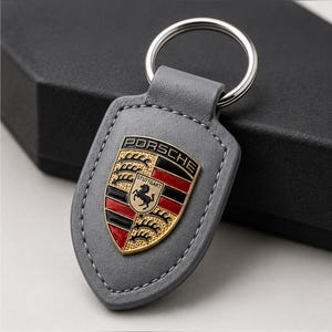 May include: A gray leather keychain featuring a shield-shaped Porsche emblem with black, red, and gold colors. The emblem displays the word PORSCHE at the top and STUTTGART in the center above a black horse. A silver metal ring is attached to the top.