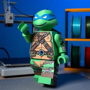 May include: A large, green, blocky plastic figure resembling a Teenage Mutant Ninja Turtle character. The figure features a blue eye mask, a tan chest plate with brown straps, and a belt with a blue L emblem. It has articulated limbs and hands.