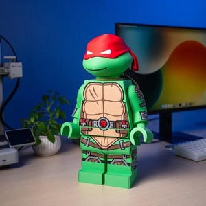 Può includere: Una grande figura in plastica verde che ricorda una Tartaruga Ninja con una maschera rossa sugli occhi. La figura presenta un pettorale color marrone chiaro, una cintura marrone con un emblema circolare rosso con una R e cinghie marroni su braccia e gambe.