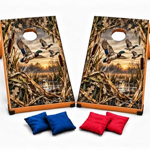 May include: Two wooden cornhole boards featuring a printed graphic of mallard ducks flying over a marsh at sunset, framed by cattails and dry grass. Each board has a circular hole at the top. Includes two blue and two red fabric bean bags.