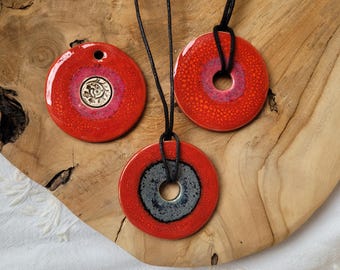 May include: Three ceramic pendant necklaces. Each pendant is a red circle with a central design and a black cord. One pendant has a silver design, another has a pink centre, and the third has a grey centre.