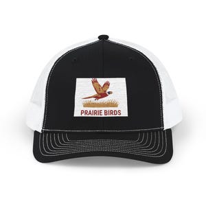 May include: A black and white trucker hat featuring a rectangular patch on the front. The patch displays an embroidered illustration of a pheasant in flight above tall grass, with the text PRAIRIE BIRDS printed in red capital letters below.