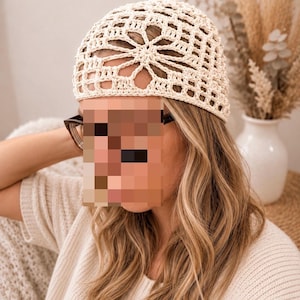 May include: A cream-colored crochet beanie featuring an intricate openwork pattern with a central star-like motif on the crown. The headwear is crafted from fine yarn, showcasing a delicate, airy texture and a snug fit that covers the top of the head.