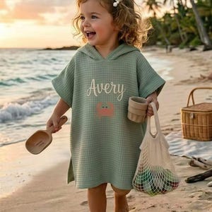 Könnte beinhalten: Ein hellgrüner Strandponcho mit Kapuze und Waffelstruktur mit dem Namen "Avery" und einer Krabben-Grafik. Das Kind hält eine Holzschaufel, einen kleinen Becher und eine Netztasche mit bunten Gegenständen. Ein Weidenkorb steht am Strand.