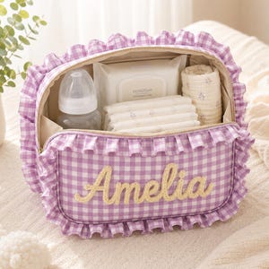 Könnte beinhalten: Eine Aufbewahrungstasche mit lila-weißem Vichy-Muster, Rüschenbesatz und dem in gelber Schreibschrift aufgestickten Namen Amelia auf der Vorderseite. Die offene Tasche enthält eine transparente Babyflasche, eine Packung Feuchttücher und weiße Tücher.