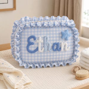 May include: A rectangular zippered pouch featuring a light blue and white gingham pattern with a ruffled border. The name Ethan is appliqued in textured blue and white yarn letters across the front, accompanied by a small blue yarn star.