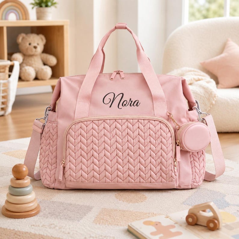 May include: A light pink diaper bag featuring a quilted chevron pattern on the front pocket, two top handles, and an adjustable shoulder strap. The name Nora is embroidered in black script on the upper front. A small round pouch is attached to the side.