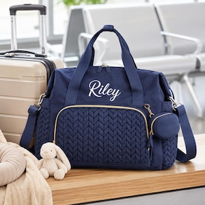 May include: A navy blue quilted travel bag featuring the name Riley embroidered in white script on the front pocket. The bag includes gold-tone zippers, two top handles, a detachable shoulder strap, and a small matching circular pouch attached.