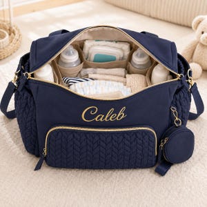 May include: A navy blue diaper bag with a quilted texture on the front pocket and sides. The name Caleb is embroidered in gold thread on the front. The bag is open, revealing internal compartments filled with baby bottles, cloths, and diapers.