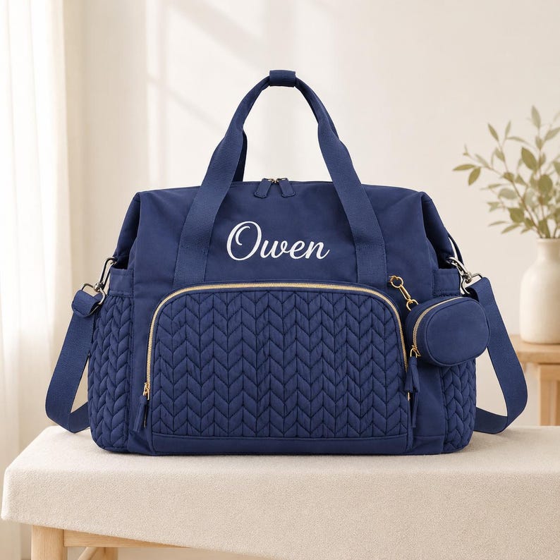 May include: A navy blue diaper bag featuring a quilted chevron pattern on the front pocket and sides. The bag has two top handles, a detachable shoulder strap, gold-tone zippers, and a small matching pouch. The name Owen is printed in white.