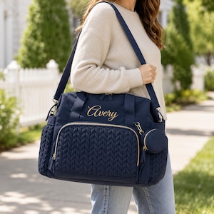 May include: A navy blue diaper bag featuring a quilted front pocket, gold-tone zippers, and the name Avery embroidered in gold script. The bag includes a matching circular pouch attached to the side and a long, adjustable shoulder strap.