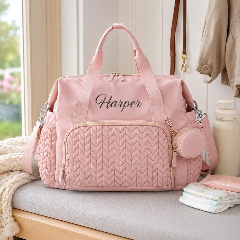 May include: A light pink diaper bag featuring a quilted herringbone pattern on the front pocket, two top handles, and a detachable circular pouch. The name Harper is embroidered in dark thread on the upper front section of the bag.