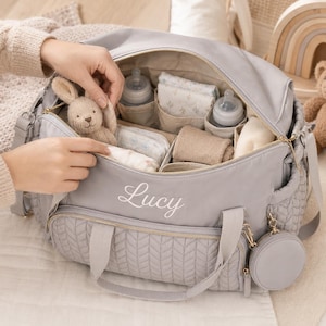 May include: A light gray diaper bag with a quilted front pocket and the name Lucy embroidered in white script. The open bag contains two baby bottles, folded cloths, and a small knitted rabbit toy. A matching circular pouch hangs from the side.